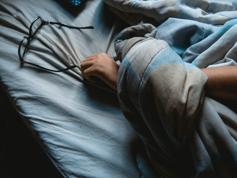 Adult resting under a blanket with a thermometer, tissues, water, and warm indoor lighting.
